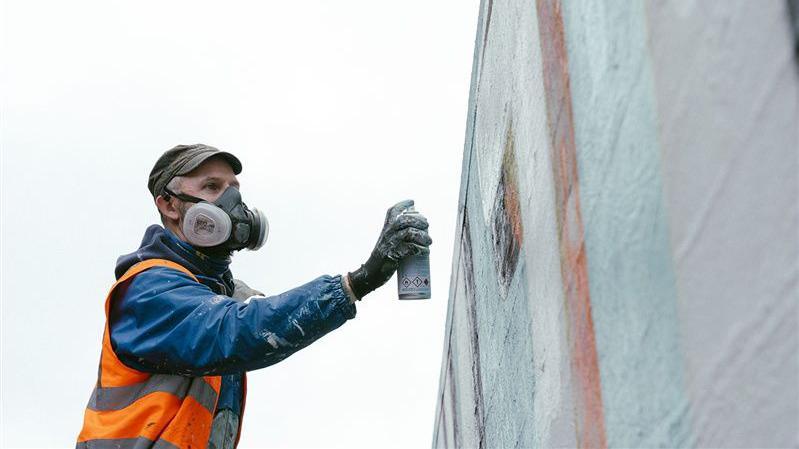 A man wearing a blue jacket, orange safety vest, and gloves is using a spray paint can to paint a large vertical surface. He is also wearing a respirator mask, and the wall being painted appears to have light-coloured sections with some geometric shapes.
