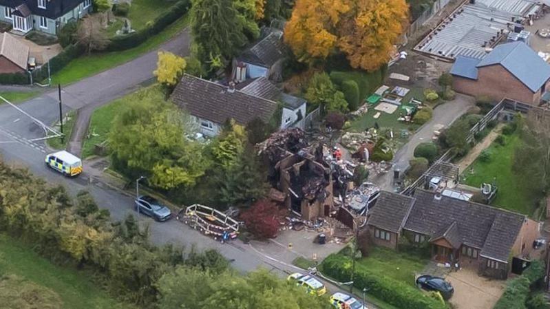 A drone view of a destroyed house with emergency vehicles parked outside and debris scattered in the garden. Nearby homes appear undamaged.