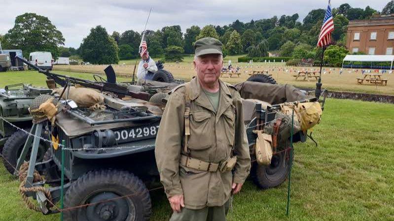 A man in green and brown military clothing is standing in front of a military vehicle that is covered in brown sandbags and black guns. He is wearing a green hat and there are American flags on the vehicle.