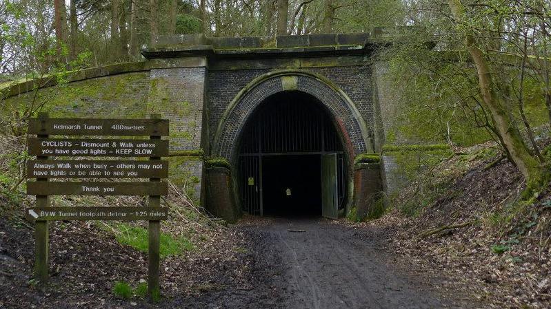 Entrance to tunnel at which iron gates are just visible. There is a grey stone archway over the entrance. There is a steep grass slope to the left and a steep mud slope to the right. A sign to the left has instructions for walkers and cyclists.