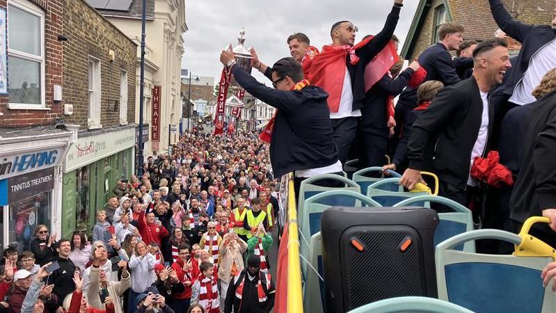 A person holding a silver trophy on an open-top bus. There are hundreds of people in the street.
