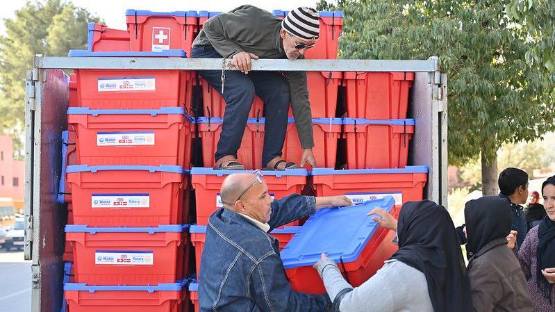 People unloading red and blue plastic boxes from a distribution truck.