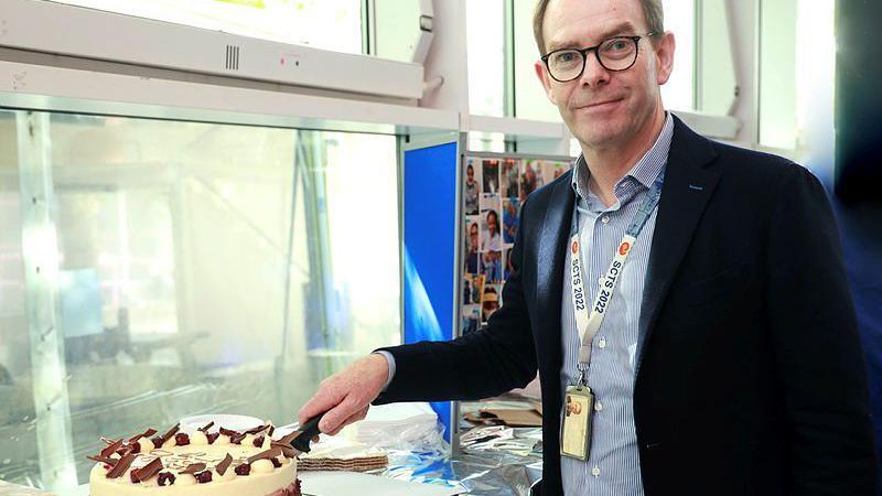 A man wearing a blue shirt and dark jacket with a lanyard around his neck uses his right arm to cut a cake with a knife. He has receding dark hair and wear glasses.