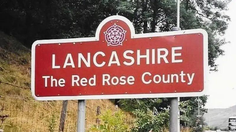 A sign for Lancashire featuring the county's red rose symbol, and the words 'The Red Rose County' underneath. Trees and a grass verge can be seen behind it.