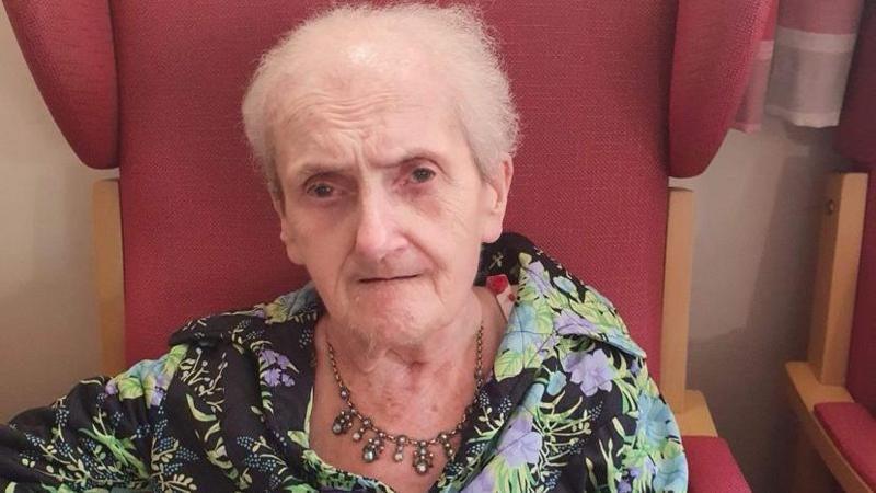 A woman with short grey hair and wearing a blouse, sits on a large red chair. She is also wearing a necklace.