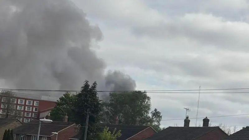 A large quantity of black smoke can be seen above the tops of houses, trees and a hotel in Cambridge. It is cloudy, with some pockets of blue sky.