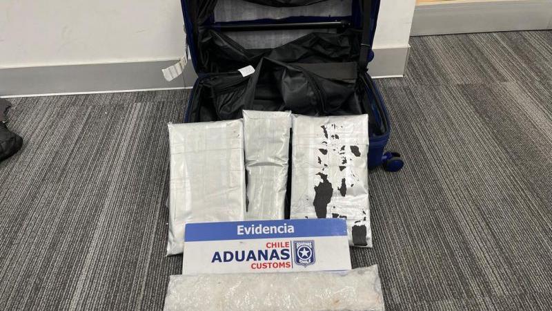 A cabin-sized suitcase which has been cut to reveal a secret compartment pictured next to bags of white powder which is methamphetamine. There is a sign by the bags of powder which says 'Evidencia Chile Aduanas Customs'.