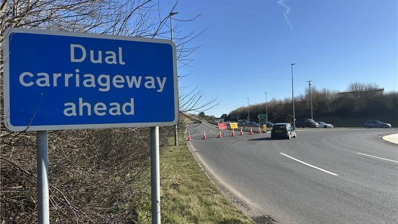 The eastbound carriageway of the A38 is closed following a crash near the Twelvewoods roundabout in Dobwalls, Cornwall. A line of traffic cones have been placed along the road of one of the roundabout exits. Signs saying the road is closed and a diversion has been set up have been erected. A blue highways sign saying "Dual carriageway ahead" is in the foreground. Several vehicles are entering the roundabout and a black Ford car is driving around the bend.