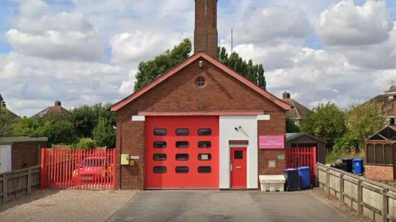 Leverton fire station. A red-brick building, set back from the main road. A red roller shutter door takes up about half of the frontage of the building. There is a space for a fire service vehicle to the left of the building behind a fence.