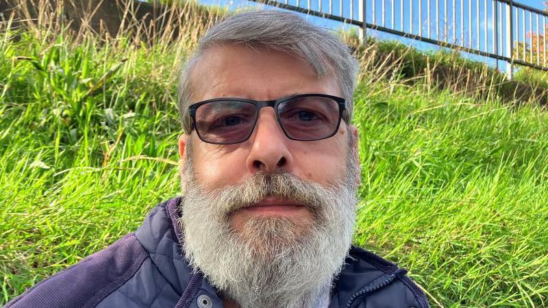 Russell Dodgson looks directly into the camera. He has short grey hair and a grey beard, is wearing dark glasses and a blue jacket and is standing in front of a grassy bank.