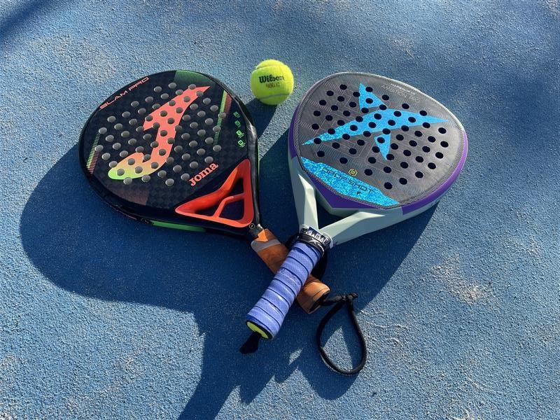 A pair of padel bats lay on the blue tarmacked ground. Between them is a green tennis ball. The racquets are similar to those used in tennis, but have a solid centre.