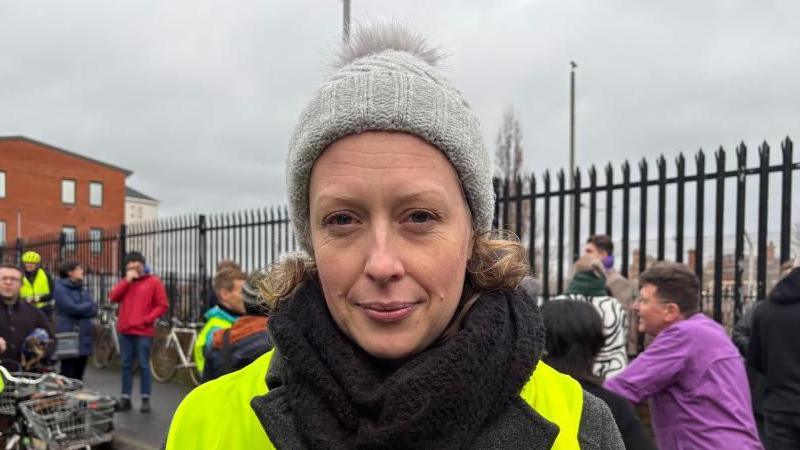 Meghan is wearing a grey woolly hat, she has a black scarf on and is wearing a high vis jacket. 