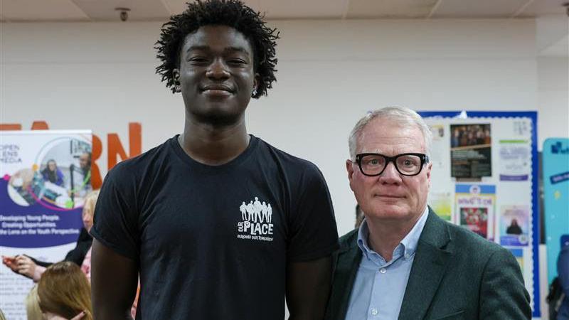 A man wearing a t-shirt which says 'The Place' on it smiles at the camera. Next to him another man wearing glasses and a blue shirt and jacket is looking at the camera. There are posters behind them in a room with white walls. 