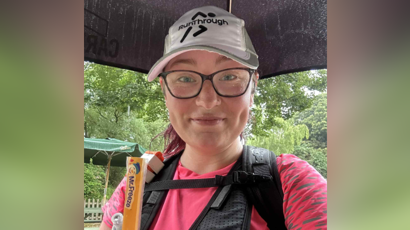 Asher Cooper is standing outside in the rain underneath an umberella. She is wearing a hot pink running top with a black running vest over the top and a white baseball cap. 