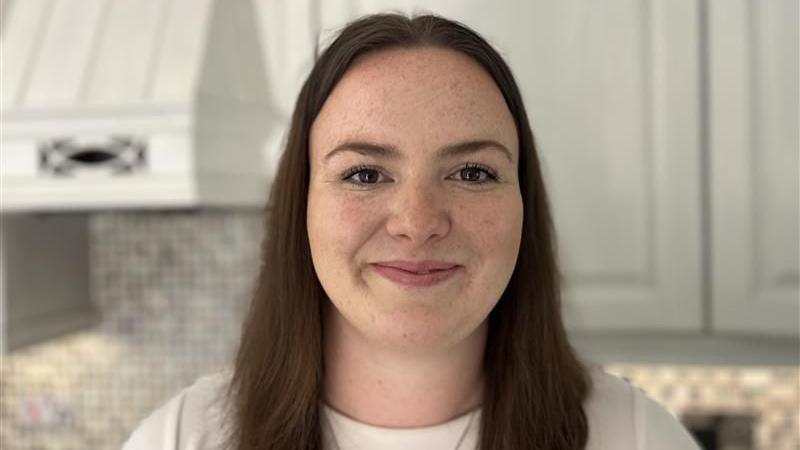 Caitlin Morgan who has long dark hair and freckles. She is wearing a white t-shirt and smiling while looking at the camera.