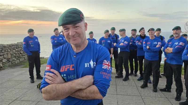 A man is smiling at the camera with his arms crossed. A group of men stand in the background behind him are smiling, laughing and chatting. They are at Land's End. The sea is in the background.