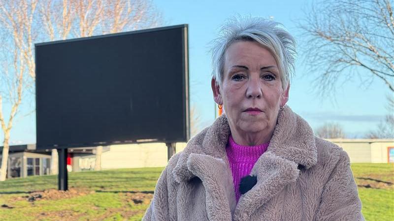 Lynn Appleby standing in front of the sign on the roundabout. She has spiky grey hair and is wearing a beige faux fur jacket.