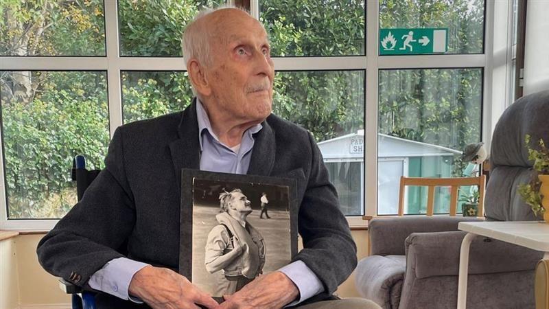 John 'paddy' hemingway in 2023. He is in a wheelchair, wearing a navy jacket and blue shirt. He is holding a black and white picture of himself taken during World War Two