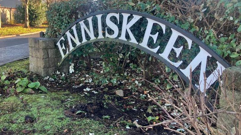A sign displaying the name Enniskeen. It is in front of a green hedge and there are stone pillars either side