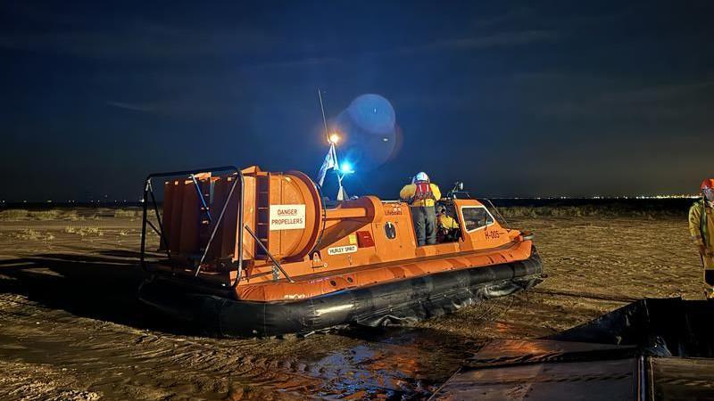 A orange RNLI hovercraft sits on the banks of the coast with the incoming tide coming towards it. One crew member is on board looking out into the sea in the dark