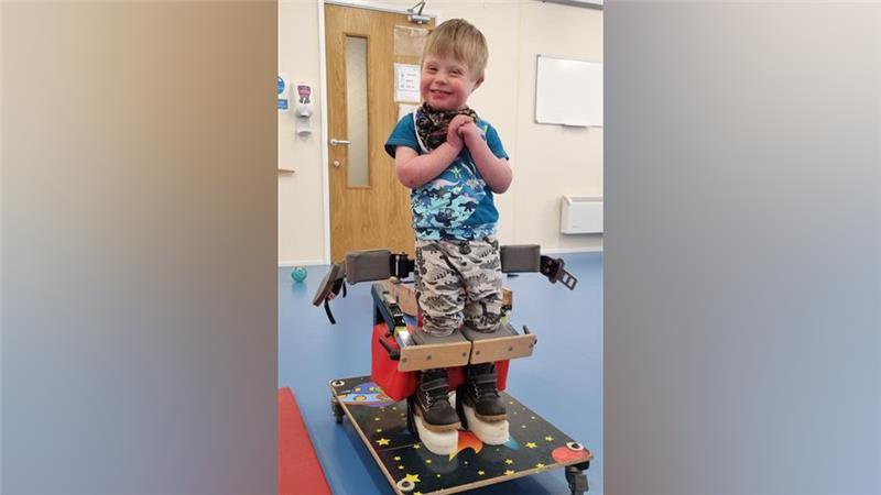 A young boy with dark blonde hair is wearing a blue camo print t-shirt and grey camo-print trousers. He is standing in a specialised standing frame which is on wheels, with his feet on blocks. He has wooden blocks in front of his knees and there is a waist strap which is undone behind him.