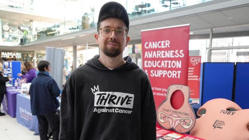 Toby Freeman, standing by his charity stall within The Forum in Norwich. He is wearing a black hoodie with a logo stating "Thrive against cancer" and wearing a black baseball cap. He has a full beard and moustache and wears glasses.