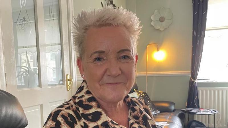 A woman with short blond hair and wearing a leopard print top sits smiling on a sofa.