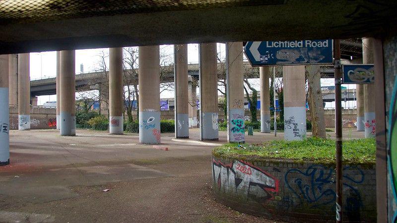 The huge beams underneath Spaghetti Junction. There is some graffiti and a Lichfield Road sign. 