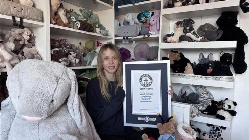 a young girl with long blonde hair holding a framed guinness world record certificate, sitting in the middle of lots of shelves of cuddly toys