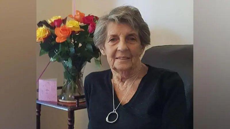 Margaret Carter sits on a chair next to a vase with yellow, orange and red roses. She has short grey hair and wears a black v-neck top with a silver necklace. 