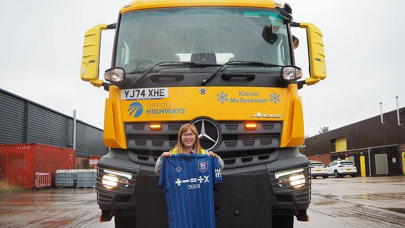 A picture of a yellow gritter vehicle with the name Kieran McSpreader written on its front. A young girl is pictured standing in front of the vehicle holding an Ipswich Town football shirt.