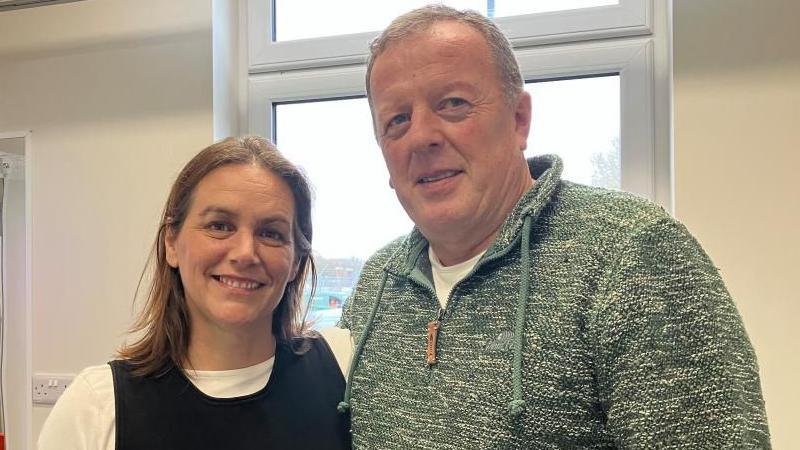 Richard and Andrea stand shoulder to shoulder. Richard has short grey hair, and wears a light green jumper and Andrea has shoulder length brown hair and wears a white t-shirt with a black knitted vest over the top.