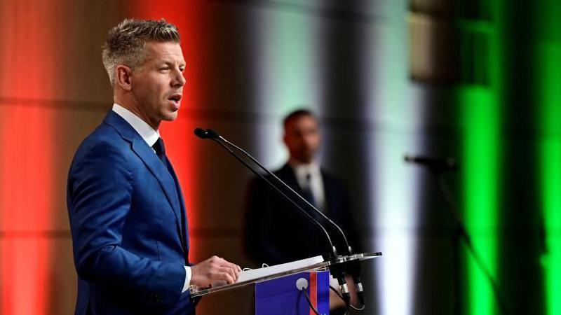 Magya speaking at a lectern with colourful lights behind him, that are red, white and green.