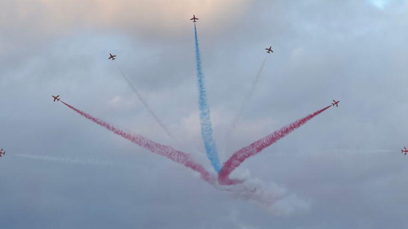 The Red Arrows flying in Guernsey. There are nine aircraft flying in the sky with red orange and blue smoke. 