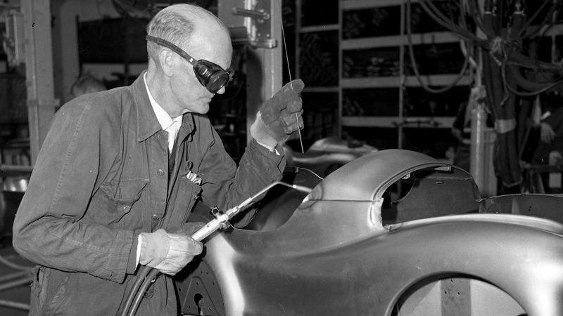 A black and white photo of a man in a boilersuit, wearing protective gloves and goggles. In his left hand he is holding a length of wire, and in his right hand is a soldering iron.