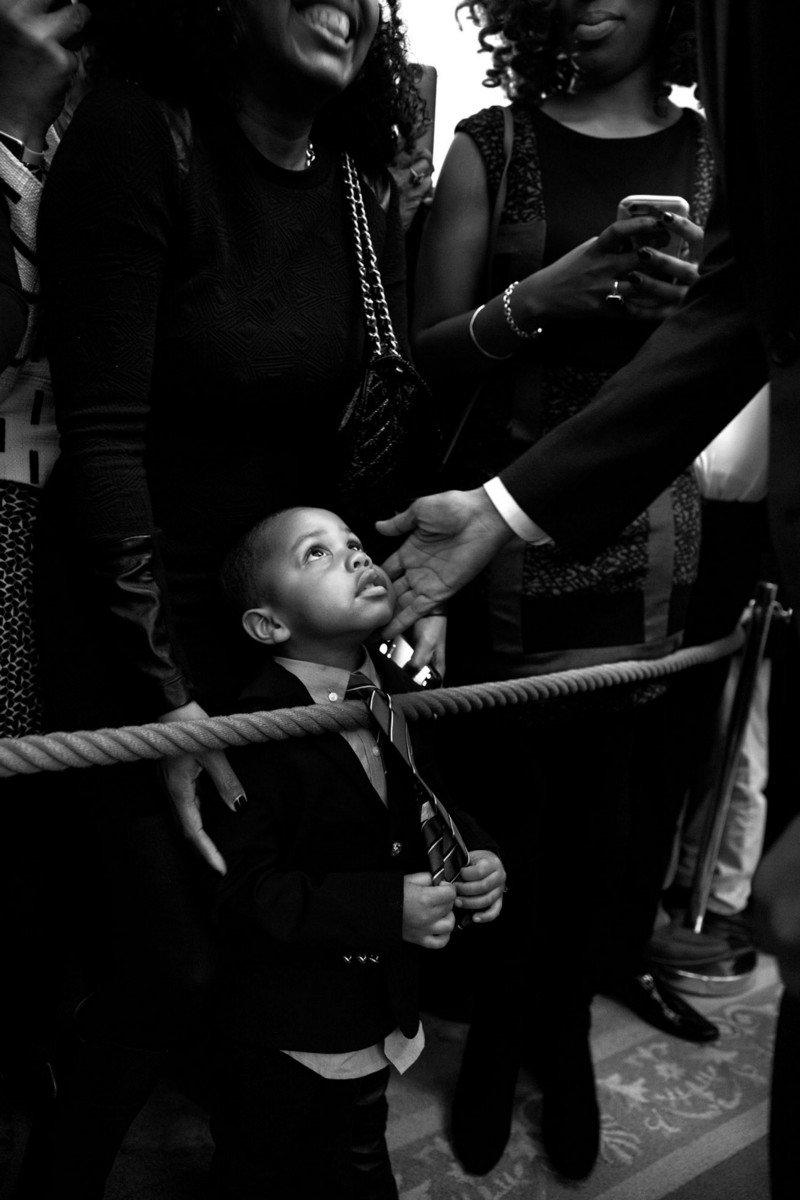 A young child dressed in a suit looks up at the unseen face of the president, as Obama reaches down and his mother stands behind him