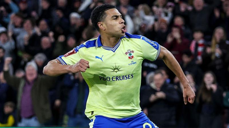 Shea Charles celebrates after scoring a goal. He is wearing a yellow football short and blue shorts. The crowd behind him celebrates.