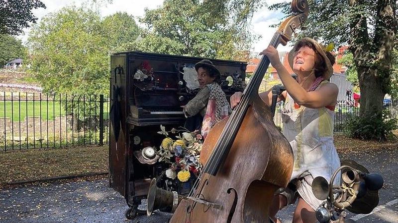 Bicycle piano and double bass duo. The pianist is turning round and looking at the camera and behind him are railings and a park. The double bass player is playing and looking up. 