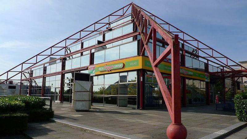 The Point seen from one corner, with its red pyramid metal bars over the corners of the building. Mirrored glass blocks make up the outer walls and there is a yellow sign over one corner saying Pizza Company. There are three storeys like a square wedding cake, with each smaller than the tier below.