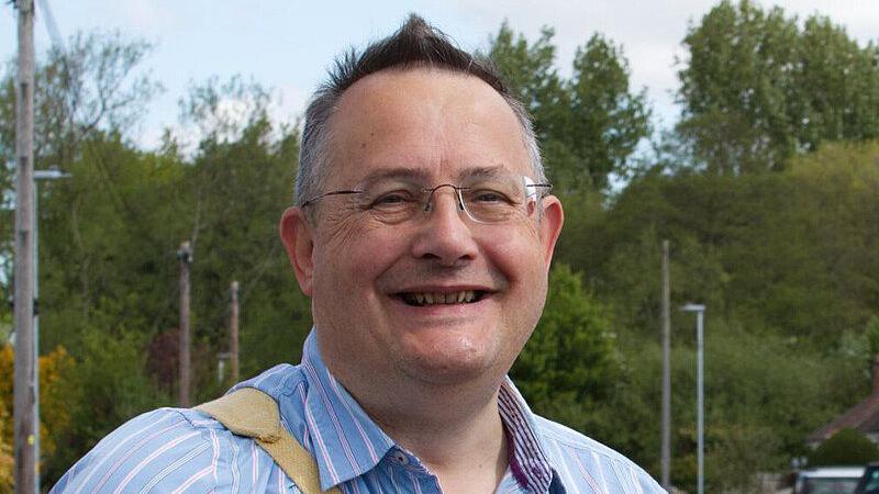 Councillor Paul Sample wearing a striped blue collared shirt and clear framed glasses, smiling at the camera. In the background there are various streetlights and a large wooded area.