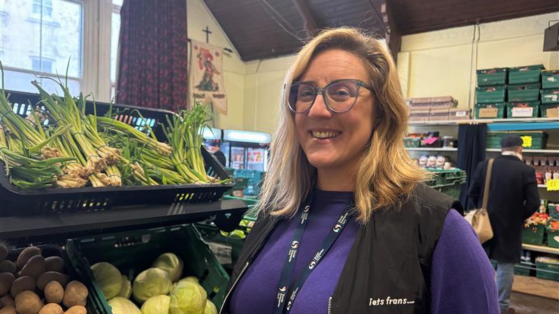 Social Enterprise Kent food projects manager Natasha Hart, wearing a purple jumper and black gilet, stood in front of green boxes of vegetables in the social supermarket. The supermarket in the background includes a man looking at produce, fridges and more boxes of food.