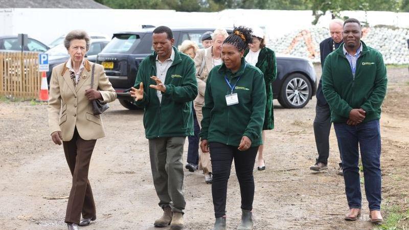 A group of six people are walking together along a dirt path in an outdoor setting, possibly a farm or rural site. In the foreground, Princess Anne is dressed smartly in a beige blazer and brown trousers, carrying a handbag and walks beside David and Brenda, wearing green jackets with name badges. David is speaking and gesturing with his hands. Behind them, three more people follow at a short distance. Parked nearby are a black SUV and another vehicle, with fencing and construction materials visible in the background. 