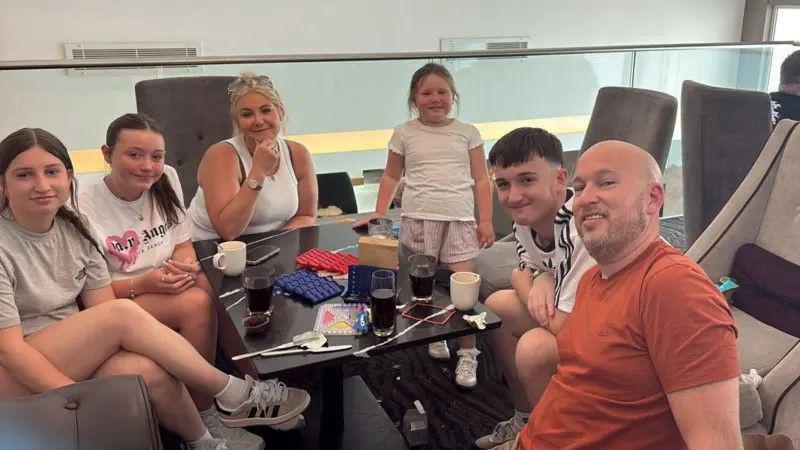 Two young girls and one younger girl, one adult woman, a young man and a man are sitting around a table in an airport lounge, with games and drinks on the table
