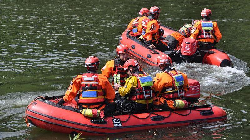 Two red search and rescue boars on a river. There are four people in each boat, wearing orange hi-vis clothing with red life jackets and red helmets.
