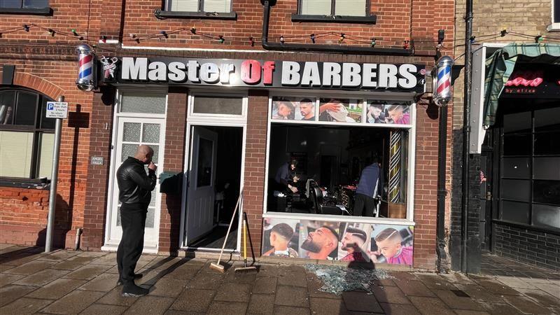A man standing outside a barber shop with its front window smashed. Broken glass is on the pavement and two brooms are propped up against a wall.