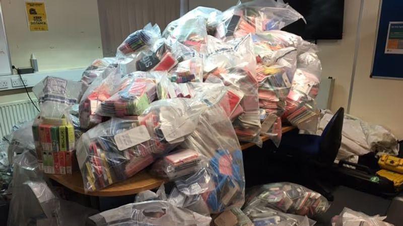 A large number of see-through plastic bags in a room and on a table. They all contain colourful vape boxes.