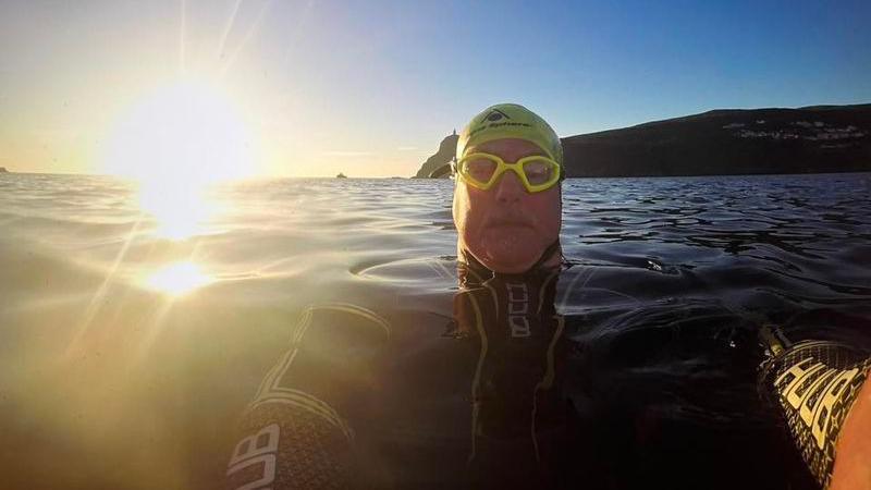 A selfie of Dave Higson in a wetsuit with goggles and a swimming cap in the sea with the sun setting behind him and a headland in the background.