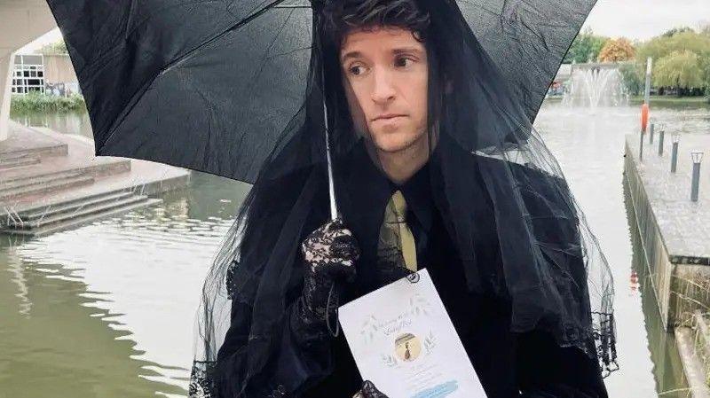 Greg James looking sad dressed in an all black mourning outfit. He's holding a black umbrella and holds an order of service for a service honouring long boi. He's near a body of water.