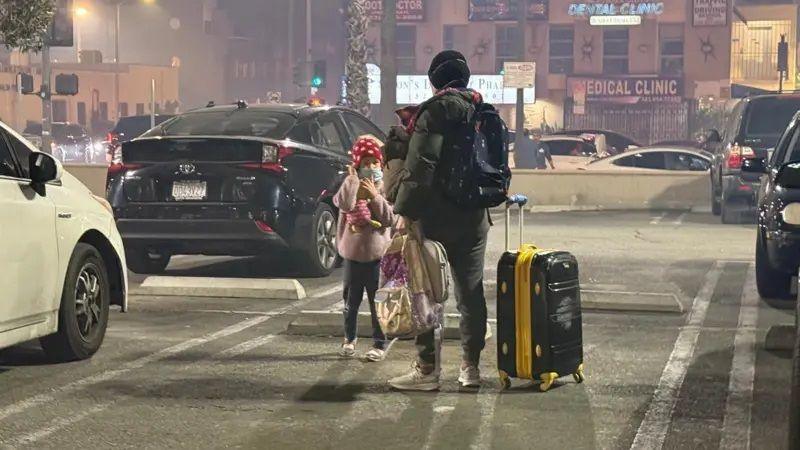 A woman with her daughter stands on a road with a suitcase in one hand and a bag in another. 