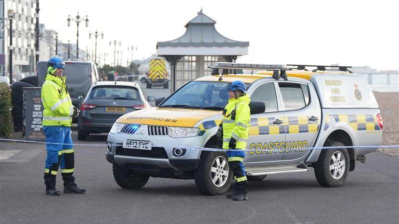 Two members of the HM Coastguard stand behind a line of tape with a HM Coastguard vehicle on the beach